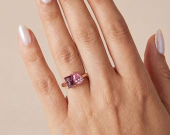 Könnte beinhalten: Ein Ring mit zwei verschiedenen Edelsteinen auf einem dünnen Metallband. Ein Stein ist ein rechteckig geschliffener violetter Edelstein, der andere ein birnenförmiger rosa Edelstein. Die Steine sind nebeneinander auf dem Ring angeordnet.