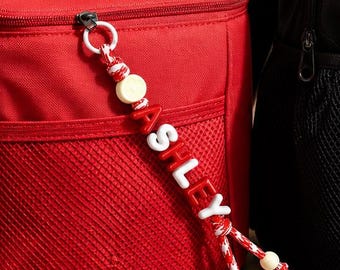 Könnte beinhalten: Eine rote Lunchtasche und ein schwarz-weißer Rucksack, jeweils mit einem personalisierten Perlen-Schlüsselanhänger. Die rote Tasche hat einen Anhänger mit der Aufschrift ASHLEY, der Rucksack einen mit CATHERINE, beide an den Reißverschlüssen.