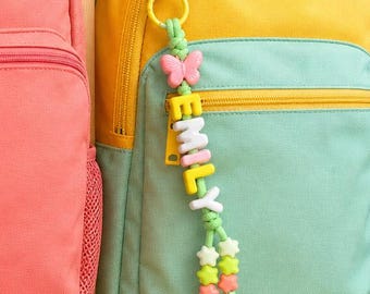 Könnte beinhalten: Drei Rucksäcke in Koralle, Gelb mit hellgrüner Tasche und Blau mit violetter Tasche. Jeder Rucksack hat einen personalisierten Perlen-Schlüsselanhänger mit Schmetterlingsanhänger und Buchstabenperlen, die die Namen AMY, EMILY und WENDY bilden.
