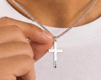 Può includere: Una collana con ciondolo a croce in metallo color argento, dotata di una catenina sottile e di un ciondolo a croce verticale. La parte inferiore della croce è decorata con tre piccole pietre preziose circolari allineate verticalmente in azzurro, rosa e blu scuro.