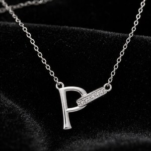 Può includere: Una collana color argento con un ciondolo a forma di lettera P. La lettera è collegata a una piccola barra rettangolare orizzontale incastonata con minuscole pietre trasparenti. Il ciondolo pende da una sottile e delicata catenina color argento.