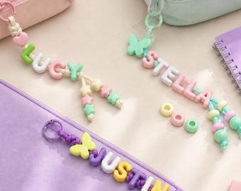 Könnte beinhalten: Drei personalisierte Reißverschlussanhänger mit bunten Kunststoff-Buchstabenperlen, die LUCY, STELLA und JUSTINA buchstabieren. Jeder Anhänger enthält einen kleinen Charm, wie einen Schmetterling oder ein Herz, und sternförmige Perlen. Ein Holzlineal zeigt 15 cm.