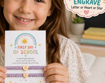 Könnte beinhalten: Zwei violette geflochtene Armbänder mit goldfarbenen Herzanhängern, graviert mit den Buchstaben M und E, befestigt an einer weißen Karte mit Regenbogenmotiv und dem Text FIRST DAY OF SCHOOL sowie einem wolkenförmigen Schild mit FREE ENGRAVE.