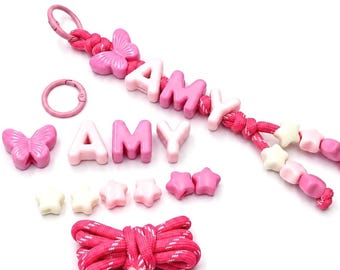 Könnte beinhalten: Ein rosa Schlüsselanhänger mit Schmetterlingsanhänger und Buchstabenperlen, die AMY buchstabieren, auf einer Schnur, begleitet von losen Perlen in Schmetterlings- und Sternform sowie den Buchstaben A, M, Y, einer aufgerollten rosa Schnur und einem Ring.