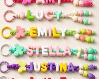 Könnte beinhalten: Acht personalisierte Schlüsselanhänger mit bunten Kunststoff-Buchstabenperlen, die AMY, LUCY, EMILY, STELLA, JUSTINA, ASHLEY, CATHERINE und WENDY buchstabieren. Jeder Anhänger hat einen Metallring, eine Schmetterlingsperle und sternförmige Perlen.