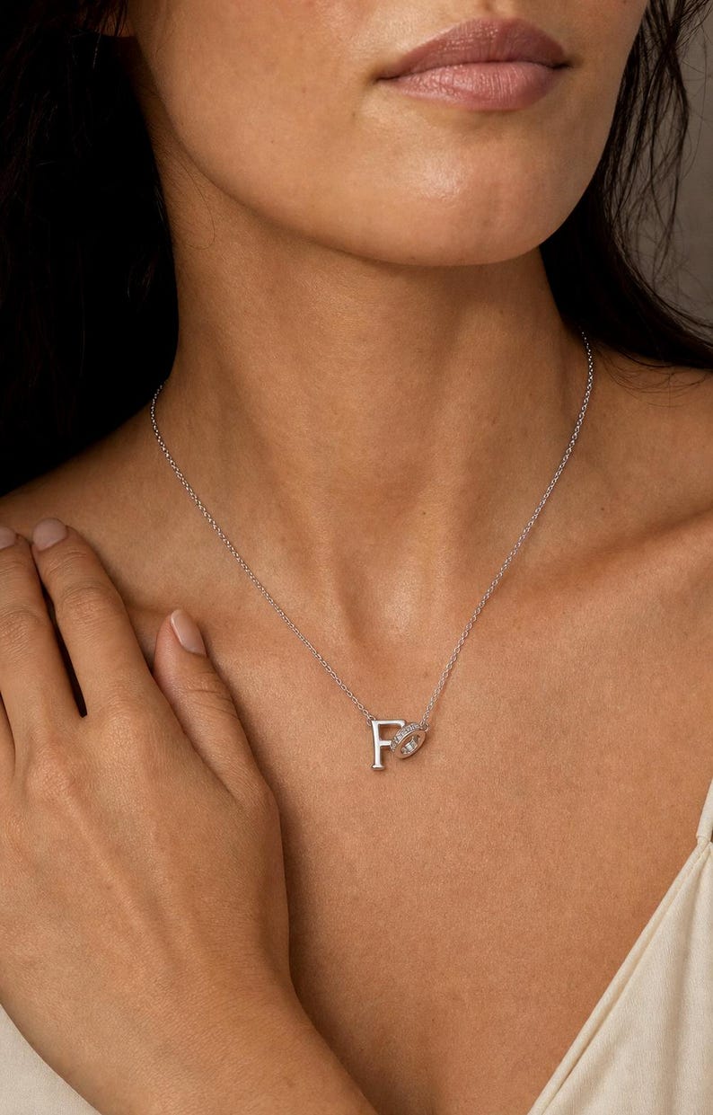 Pu&ograve; includere: Una collana color argento con un ciondolo a forma di lettera F maiuscola attaccato a un piccolo cerchio tempestato di pietre trasparenti scintillanti. Il ciondolo pende da una sottile e delicata catenina color argento al collo.