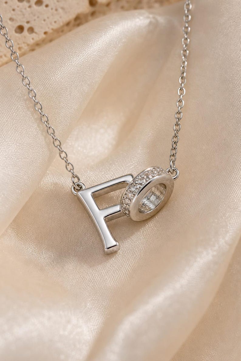 Pu&ograve; includere: Una collana color argento con un ciondolo che presenta la lettera F collegata a un elemento circolare incastonato con piccole pietre trasparenti. Il ciondolo pende da una sottile catenina color argento dall'aspetto metallico lucido.