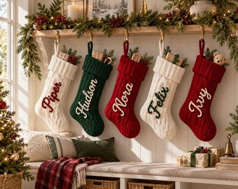 Könnte beinhalten: Fünf in einer Reihe hängende Weihnachtsstrümpfe aus Zopfstrick mit den in Kontrastfarben gestickten Namen Piper, Hudson, Nora, Felix und Ivy. Die Strümpfe sind cremefarben, dunkelgrün und rot und jeweils mit einem Rippbündchen und Tannengrün versehen.