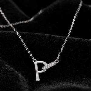 Pu&ograve; includere: Una collana color argento con un ciondolo a forma di lettera P. La lettera &egrave; collegata a una piccola barra rettangolare orizzontale incastonata con minuscole pietre trasparenti. Il ciondolo pende da una sottile e delicata catenina color argento.