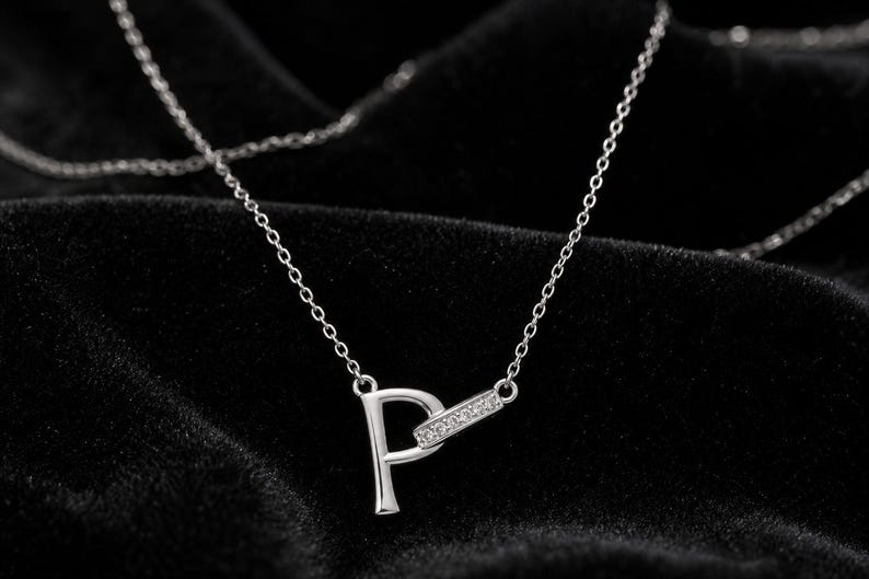 Pu&ograve; includere: Una collana color argento con un ciondolo a forma di lettera P. La lettera &egrave; collegata a una piccola barra rettangolare orizzontale incastonata con minuscole pietre trasparenti. Il ciondolo pende da una sottile e delicata catenina color argento.