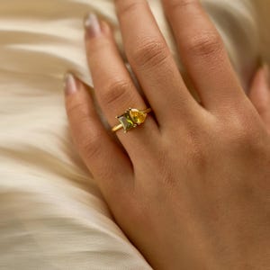 Peut inclure: Une bague en or ornée de deux pierres précieuses distinctes : une pierre rectangulaire vert olive et une pierre jaune en forme de poire. Les pierres sont serties côte à côte sur un anneau en or fin et poli, créant un design asymétrique.