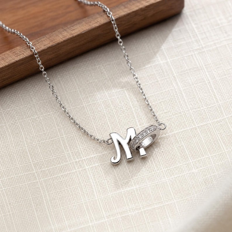 Pu&ograve; includere: Una collana color argento con una catenina sottile e un ciondolo composto da una lettera M stilizzata collegata a un piccolo cerchio decorato con minuscole pietre trasparenti. I componenti in metallo presentano una finitura lucida e brillante.