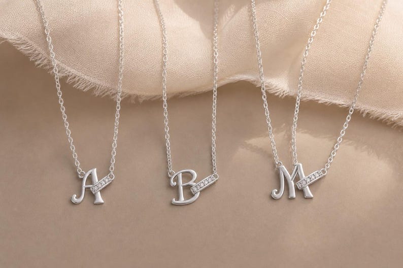 Pu&ograve; includere: Tre collane color argento con le lettere A, B e M come ciondoli. Ogni lettera &egrave; attaccata a una sottile catena d'argento ed &egrave; accentuata da una piccola barra rettangolare incastonata con minuscole pietre chiare e scintillanti posizionate in diagonale.