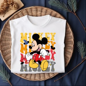 May include: White t-shirt featuring a graphic of Mickey Mouse in a running pose. The design includes the word "Mickey" in yellow, red, and gray, with star accents. The shirt has a classic crew neck.