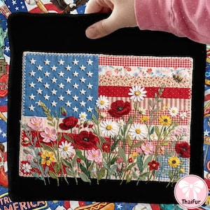 Könnte beinhalten: Ein handgefertigtes Textilkunstwerk mit einem Design der amerikanischen Flagge. Die Flagge enthält gestickte Sterne, Streifen und eine Blumenwiese mit Mohnblumen, Gänseblümchen und anderen Blumen. Das Kunstwerk ist vor einem schwarzen Hintergrund.