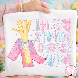 Könnte beinhalten: Weißes Sweatshirt mit einer skurrilen Grafik eines Käse-Sticks in einer rosa Jacke, die mit einer blauen Schleife gebunden ist. Der Text "in my string cheese era" ist in rosa und blauen Buchstaben.