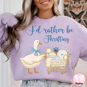 May include: A light purple sweatshirt with the text "I'd rather be Thrifting" in blue script. A cartoon goose with a blue bow pulls a shopping cart filled with vintage items, including dishes and a lamp.