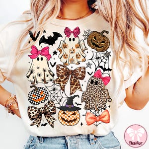 May include: Cream-colored Halloween-themed t-shirt featuring various ghost designs with bows, pumpkins, spiders, and bats. The ghosts have different patterns, including leopard print and pumpkin faces. The shirt has a playful and festive design.