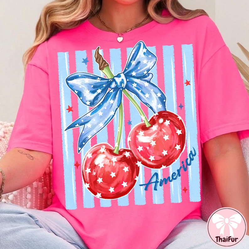 May include: A pink t-shirt featuring a patriotic design. The graphic includes two red cherries with white stars, a blue bow with white stars, and blue and white striped background. The word "America" is written in blue.