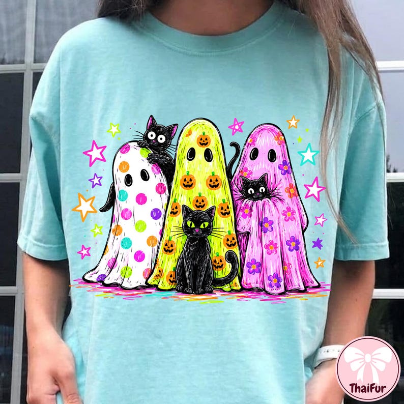 Pu&ograve; includere: Una t-shirt azzurro chiaro con una grafica di tre fantasmi e gatti neri. Un fantasma ha pois colorati, uno ha zucche arancioni e uno ha fiori viola. Il design include stelle colorate sparse attorno alle figure.
