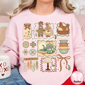 Może przedstawiać: R&oacute;żowa bluza z nadrukiem z ilustracjami w stylu wiejskim. Projekt zawiera słoik, misie, kurę w koszu, czajnik, miski, lampę i kokardę. Og&oacute;lna estetyka jest vintage i przytulna.
