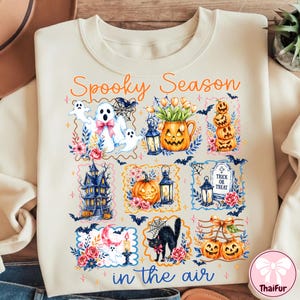 May include: A cream-colored sweatshirt featuring a graphic print with the text Spooky Season in the air. The design includes illustrations of ghosts, a jack-o'-lantern, a haunted house, a black cat, bats, flowers, and a tombstone with Trick or Treat.