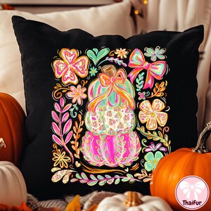 May include: Black decorative pillow featuring a stacked pumpkin design with a bow and floral accents. The pumpkins are pink, green, and white, surrounded by colorful flowers and leaves. A festive fall-themed home decor item.