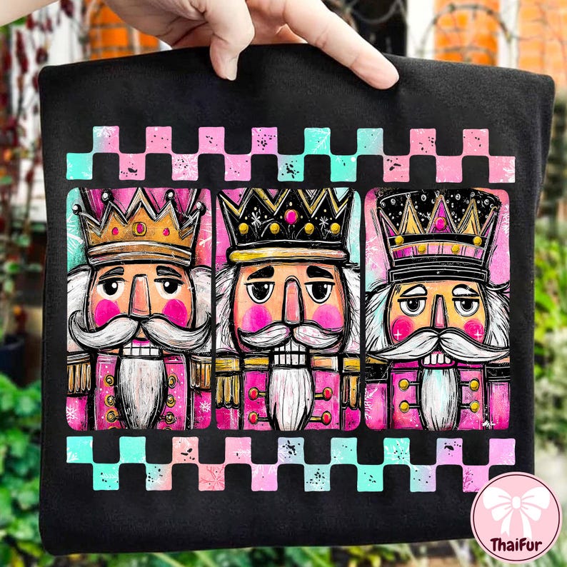 K&ouml;nnte beinhalten: Schwarzes Sweatshirt mit einem farbenfrohen Aufdruck von drei Nussknacker-Figuren mit Kronen und Schnurrb&auml;rten. Das Design enth&auml;lt rosa, goldene und t&uuml;rkisfarbene Akzente mit einem karierten Rand. Das Sweatshirt wird von einer Hand gehalten.