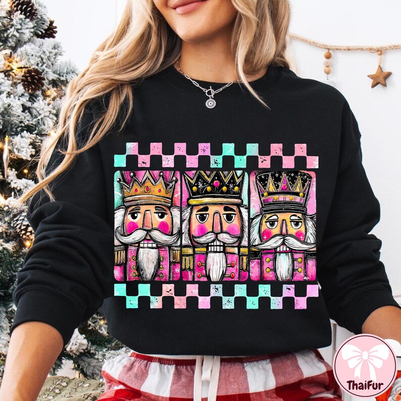 K&ouml;nnte beinhalten: Schwarzes Sweatshirt mit einem farbenfrohen Grafikdesign von drei Nussknacker-Figuren mit Kronen und Schnurrb&auml;rten. Das Design enth&auml;lt rosa, t&uuml;rkisfarbene und goldene Akzente vor einem karierten Hintergrund. Ein festliches Kleidungsst&uuml;ck zum Thema Feiertage.