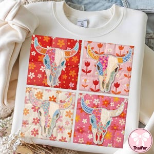 Puede incluir: Sudadera blanca con un diseño de patchwork de cuatro calaveras de vaca. Cada calavera está colocada sobre un fondo floral diferente, con las calaveras hechas de tela colorida. El diseño tiene una apariencia acolchada.