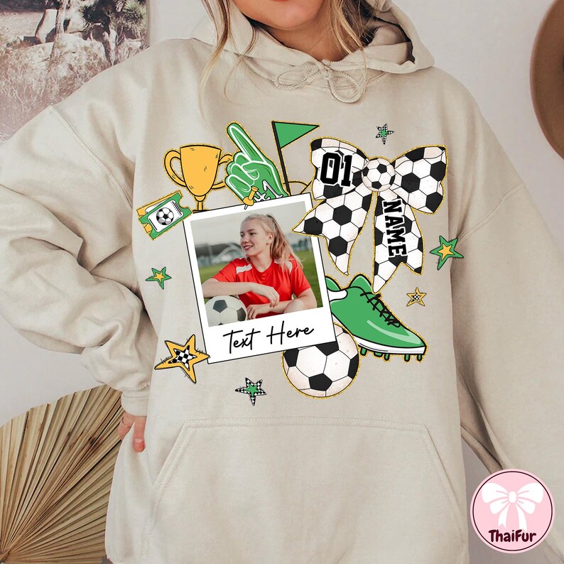 May include: A beige hoodie with a graphic design featuring a soccer ball, a trophy, a photo of a girl playing soccer, a bow with the text "NAME" and the number "01", and the text "Text Here".