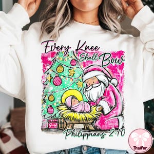 May include: White sweatshirt with a Christmas-themed graphic. The design features a pink Santa Claus kneeling before baby Jesus in a manger, with a Christmas tree and the text "Every Knee Shall Bow Philippians 2:10".