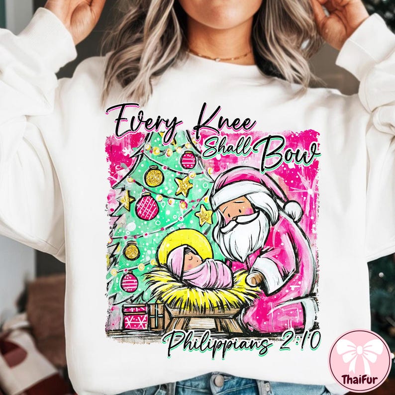 May include: White sweatshirt with a Christmas-themed graphic. The design features a pink Santa Claus kneeling before baby Jesus in a manger, with a Christmas tree and the text "Every Knee Shall Bow Philippians 2:10".