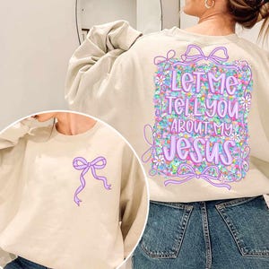 Puede incluir: Una sudadera beige con un dise&ntilde;o floral y el texto "Let Me Tell You About My Jesus" en la espalda. La parte delantera de la sudadera tiene un lazo morado.