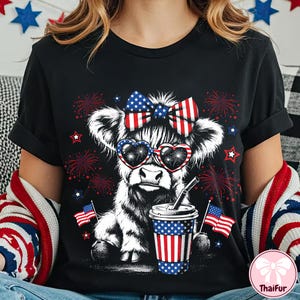 May include: Black t-shirt featuring a cow illustration with heart-shaped sunglasses and a bow, both in red, white, and blue. The cow is holding a cup with a straw and American flag accents. Fireworks and stars complete the patriotic design.
