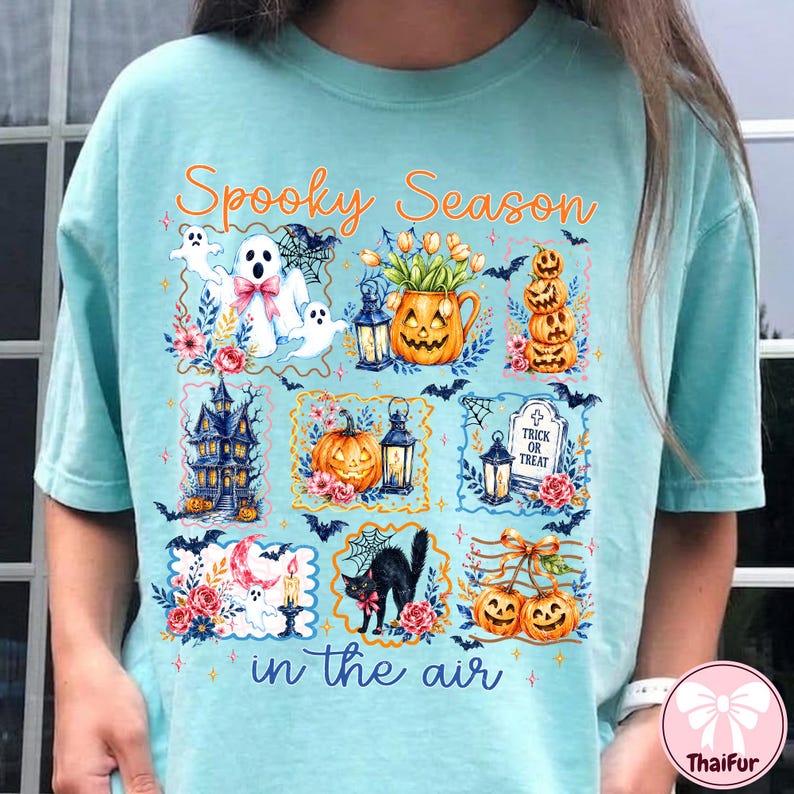 May include: A light blue t-shirt featuring the orange text Spooky Season in the air and a graphic print of Halloween elements including ghosts, pumpkins, a haunted house, a black cat, a tombstone, lanterns, and flowers in a grid layout.