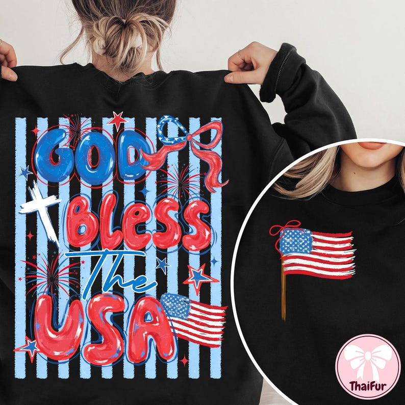 Op de afbeelding: Zwart sweatshirt met de tekst "God Bless The USA" in rode en blauwe letters. Het ontwerp bevat een kruis, vuurwerk, sterren en een Amerikaanse vlag. Een kleiner beeld van de Amerikaanse vlag staat aan de rechterkant.
