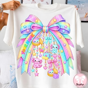 Könnte beinhalten: Ein weißes T-Shirt mit einer großen, glitzernden, regenbogenfarbenen Schleifengrafik. Das Design zeigt hängende Goldketten mit Anhängern in Form von Axolotln, Teigtaschen, einer Pfote und einem Koffer. Der Text "YOUR NAME" steht in Orange links.
