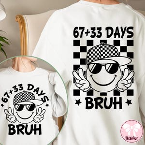 May include: White sweatshirt with a black and white graphic. The graphic features a smiley face wearing sunglasses and a checkered hat, with the text "67+33 DAYS" and "BRUH". The smiley face is making a peace sign with both hands.