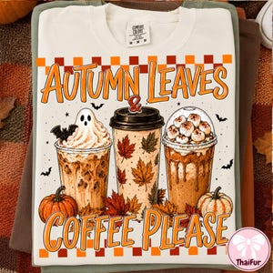Może przedstawiać: Kremowy t-shirt z grafiką przedstawiającą trzy kubki kawy, ducha, nietoperze i dynie. Napis głosi Autumn Leaves & Coffee Please. Wzór zawiera pomarańczowe i brązowe liście klonu oraz szachownicę na górnej i dolnej krawędzi nadruku.