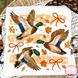May include: A white t-shirt featuring a graphic print of three mallard ducks in flight, surrounded by orange autumn maple leaves, light orange horizontal brush strokes with white polka dots, and three orange ribbon bows.