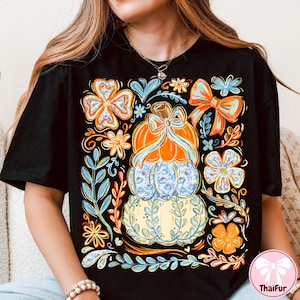 May include: Black t-shirt featuring a stacked pumpkin design with a bow, surrounded by floral and leaf accents in orange, blue, and yellow. The design includes a light blue pumpkin with a floral pattern.