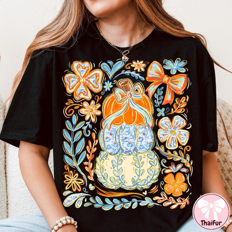 May include: Black t-shirt featuring a stacked pumpkin design with a bow, surrounded by floral and leaf accents in orange, blue, and yellow. The design includes a light blue pumpkin with a floral pattern.