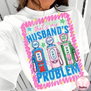 May include: White t-shirt with a colorful graphic design. The design features three vintage gas pumps in blue, pink, and green, with the text "that's my HUSBAND'S PROBLEM" in blue and pink.