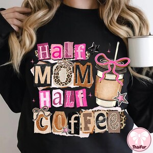 May include: Black sweatshirt with the text "Half Mom Half Coffee" in a collage style. The design includes a coffee cup with a pink bow, stars, and leopard print accents. A white mug is held in the background.