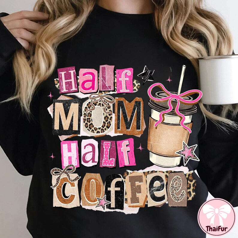 May include: Black sweatshirt with the text "Half Mom Half Coffee" in a collage style. The design includes a coffee cup with a pink bow, stars, and leopard print accents. A white mug is held in the background.