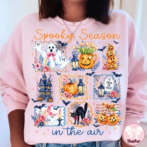 May include: A light pink sweatshirt featuring the orange text Spooky Season in the air. The graphic includes illustrations of ghosts, jack-o'-lanterns, a haunted house, a black cat, a tombstone with Trick or Treat, lanterns, bats, and flowers.