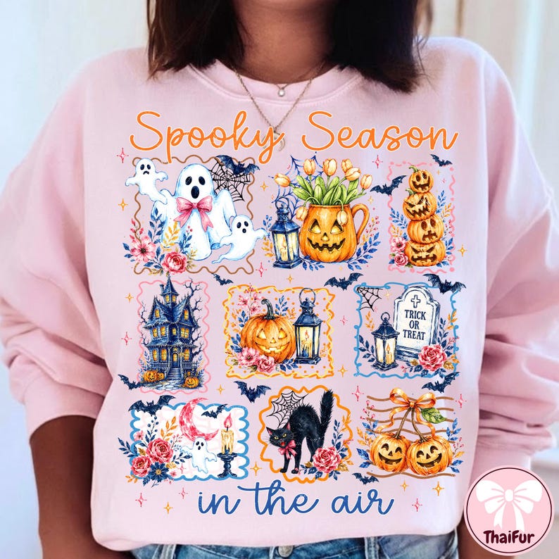 May include: A light pink sweatshirt featuring the orange text Spooky Season in the air. The graphic includes illustrations of ghosts, jack-o'-lanterns, a haunted house, a black cat, a tombstone with Trick or Treat, lanterns, bats, and flowers.