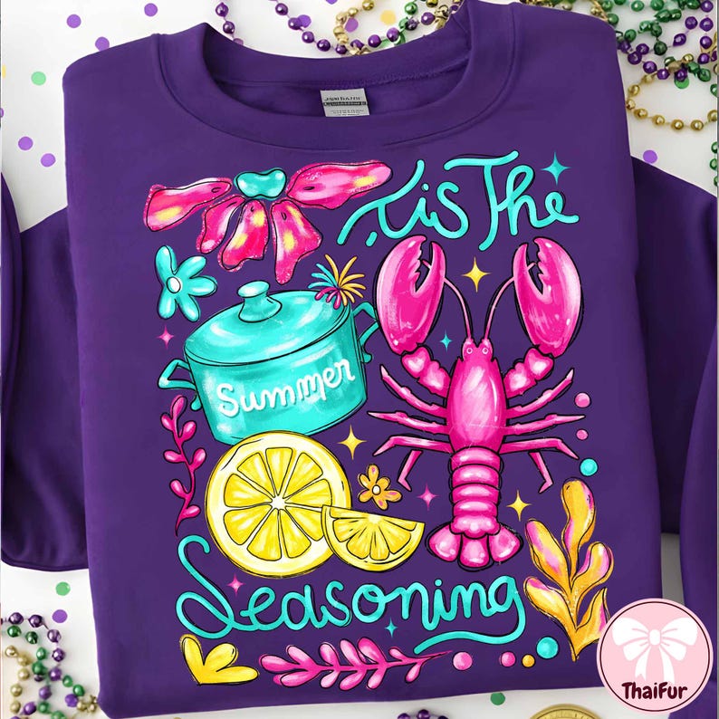 May include: A purple sweatshirt with a colorful graphic design. The design includes a pink lobster, a lemon, a teal pot with the word "Summer", and the phrase "'Tis The Seasoning". The design is surrounded by floral and decorative elements.