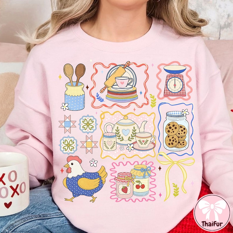 May include: A light pink sweatshirt with a colourful, illustrated design featuring kitchen and baking-themed elements. The design includes a chicken, jars of jam, a teapot, and a scale.